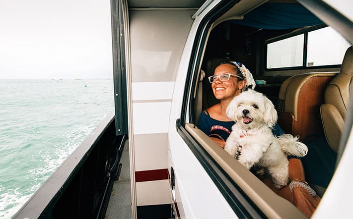 A woman and small dog looking out the window of an RV camper van smiling and looking at the ocean outside